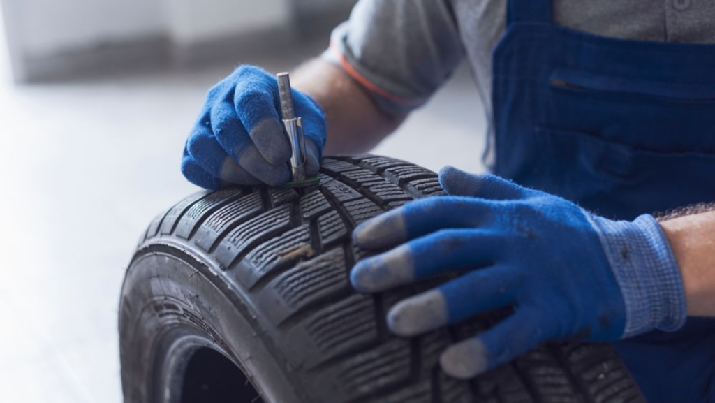 A mechanic is using a tire depth gauge to check the tread of a tire, wearing blue gloves and working in a garage setting.