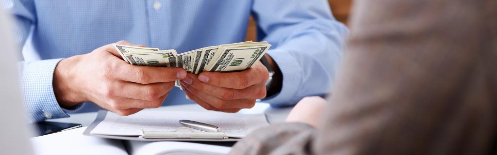 A person in a blue shirt counting U.S. dollar bills while sitting across from another person at a desk with documents and a pen visible.