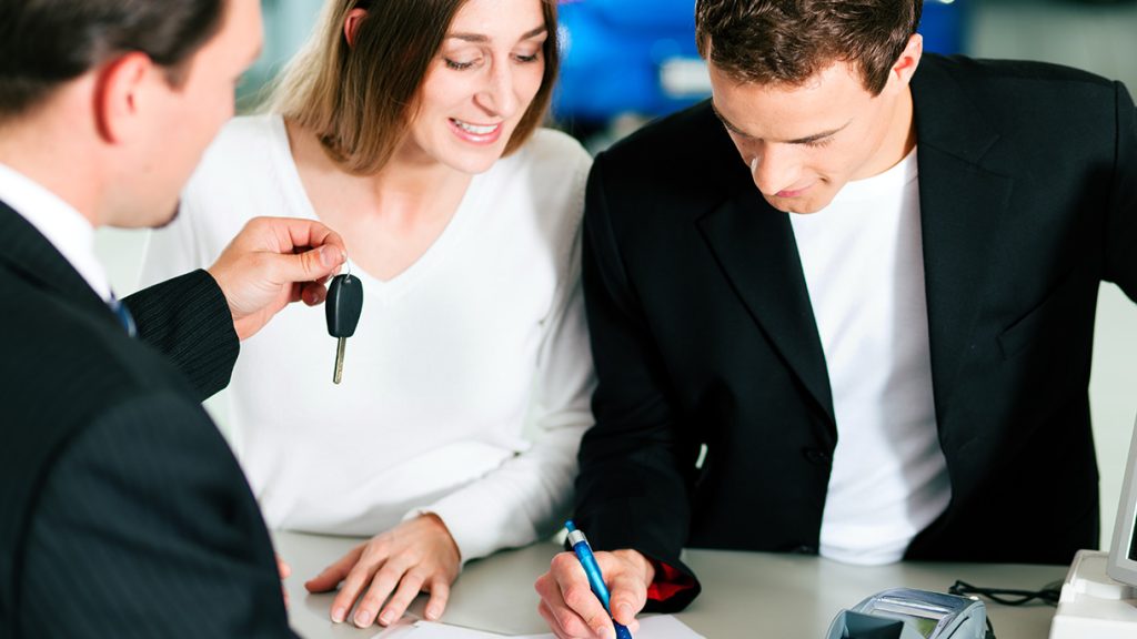 Car dealership customer signs paperwork while a salesperson hands over a vehicle key during the purchase process.