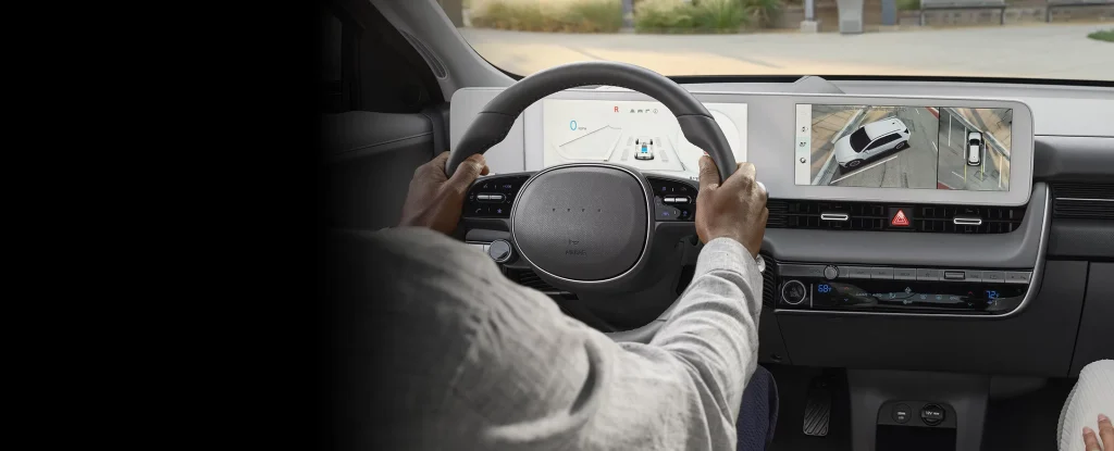 A person driving a car, holding the steering wheel, with a dashboard display showing a dual screen interface, including a map and a rearview view of the vehicle.