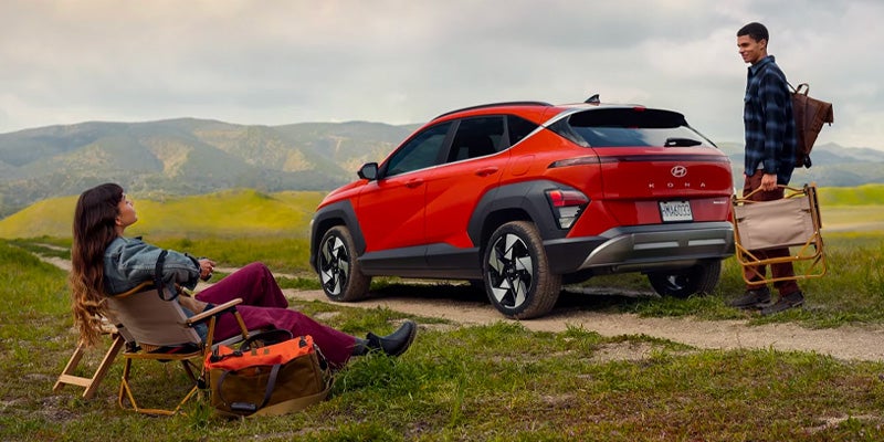 Red Hyundai Kona parked on grassy trail with a couple setting up camping chairs nearby